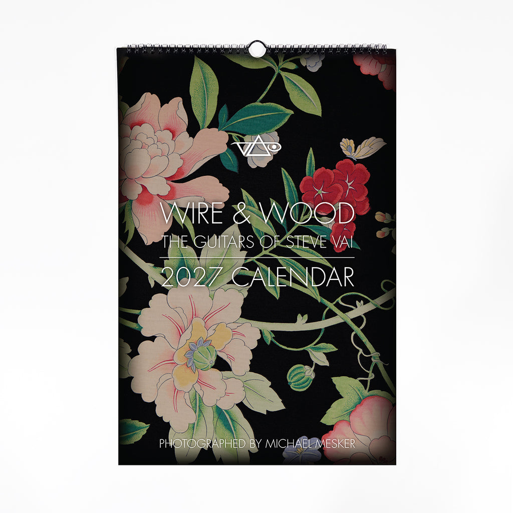 A hanging wall calendar titled 'WIRE & WOOD: THE GUITARS OF STEVE VAI 2027 CALENDAR' with a black background and large floral artwork in pink, red, cream, and green. The cover includes a spiral binding at the top and a wire hanger loop at the center.