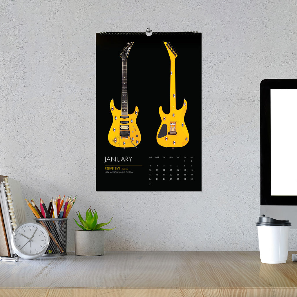 A wall calendar featuring a yellow electric guitar on a black background, hanging on a textured light gray wall above a desk with a computer monitor, clock, pencils, and a small plant.
