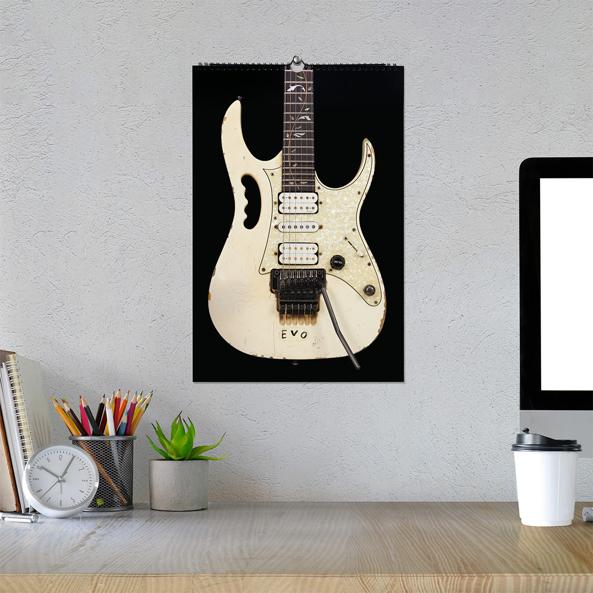 A wall calendar featuring EVO on a black background, hanging on a textured light gray wall above a desk with a computer monitor, clock, pencils, and a small plant.