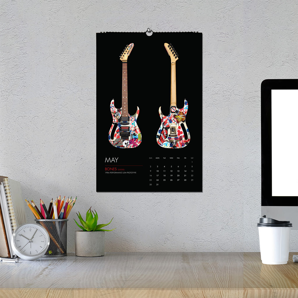 A wall calendar featuring the BONES guitar on a black background, hanging on a textured light gray wall above a desk with a computer monitor, clock, pencils, and a small plant.