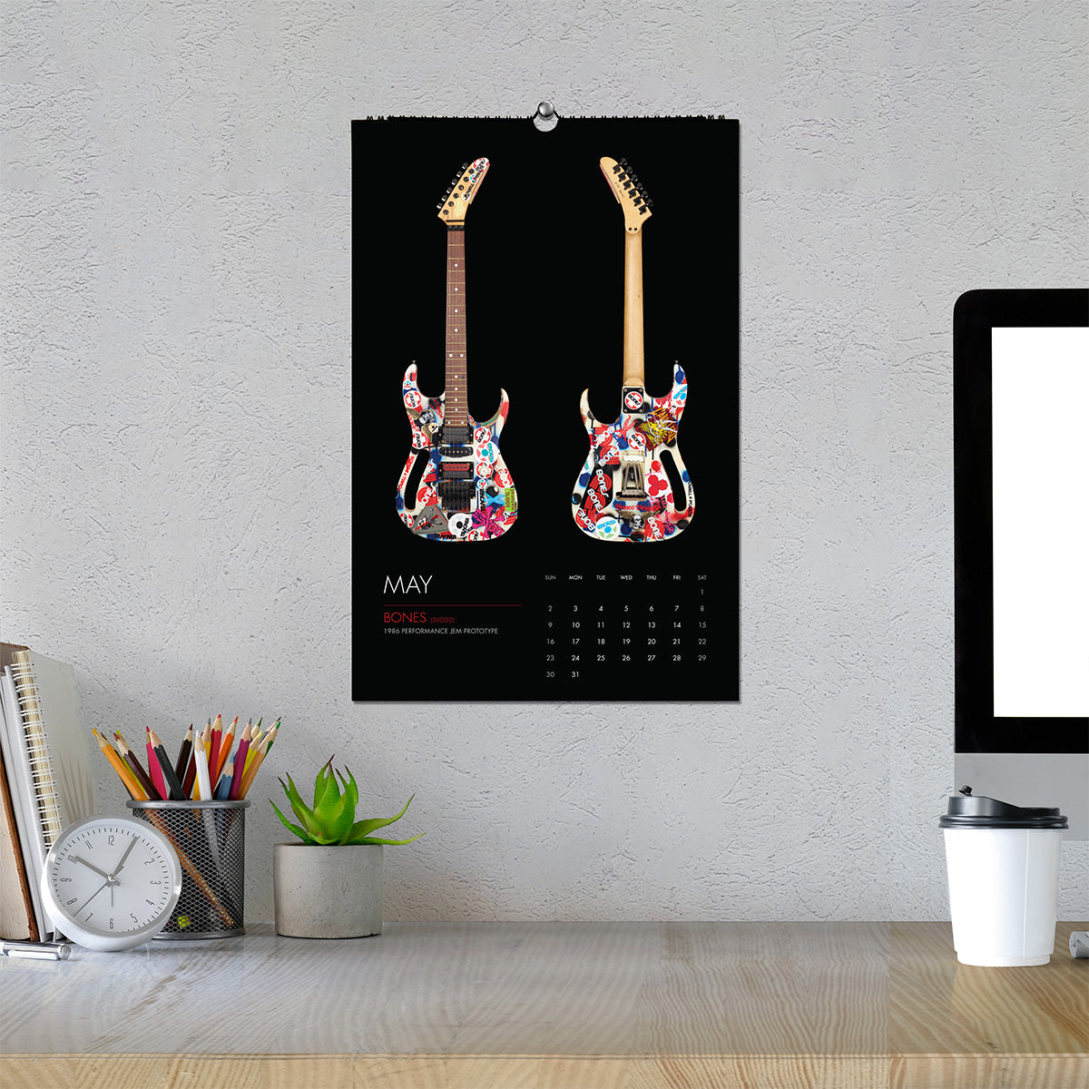 A wall calendar featuring the BONES guitar on a black background, hanging on a textured light gray wall above a desk with a computer monitor, clock, pencils, and a small plant.